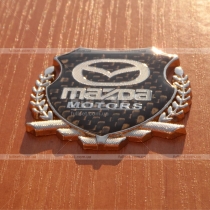 Эмблема герб Mazda на карбоновом фоне (размер: 6,5 см на 5,5 см)